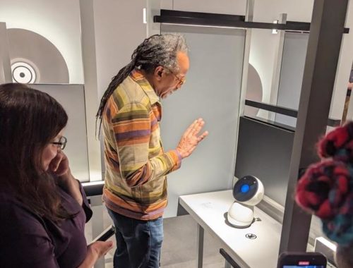 Fellow Louis Massiah waves to Jibo, a small social robot sitting on a table.