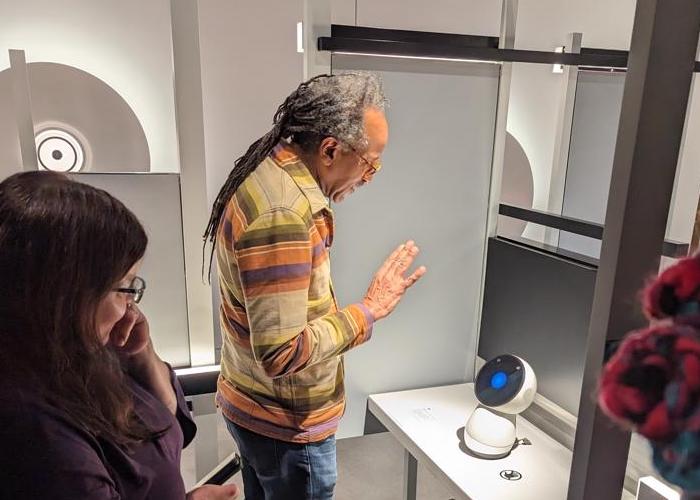 ODL Fellow Louis Massiah waves to a small white robot sitting on a table as curator Lindsey Bartholomew watches. The robot has a circular "face" with a digital screen that animates as it responds.