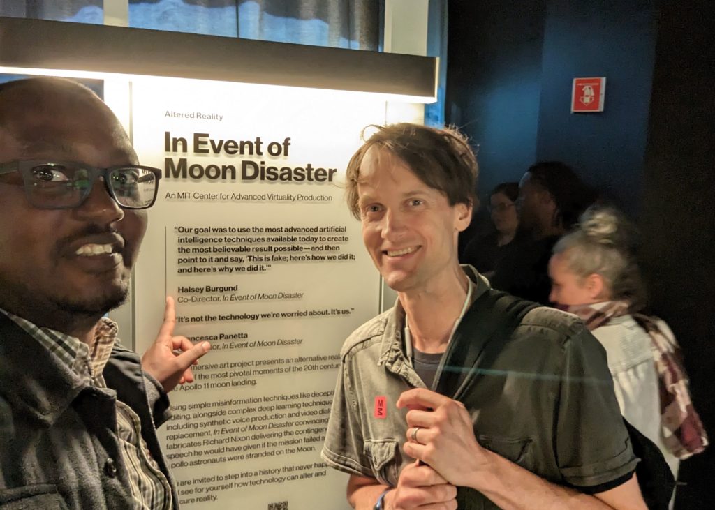 Max Musau and Halsey Burgund stand in front of a sign describing "In Event of Moon Disaster." Musau is pointing at Burgund's name where he is listed as a co-creator of the project.