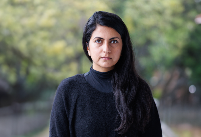 A photo of Meghna Singh from the torso up. She has long black hair and is wearing a black sweater. She is standing in front of a blurred background of plants outside.