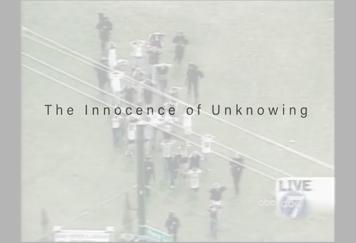A screenshot of a news broadcast showing small figures holding their hands up as they walk away from a building. The words "The Innocence of Unknowing" are written in black text on top of the image.