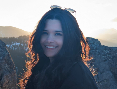 [Image Description: Nouf Aljowaysir, an Arab woman with long, dark, curly hair, smiles warmly at the camera. She is standing outside in nature, framed by snow and a mountainous backdrop.]